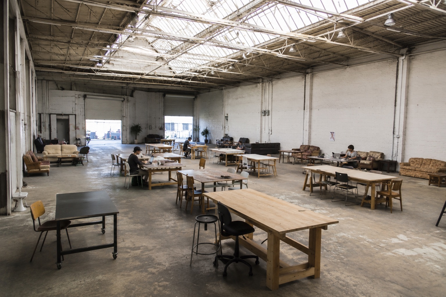 A white-brick warehouse under skylights: long wooden tables with people working at them, mismatched second-hand sofas around the edges, roll-up doors open at the far end.